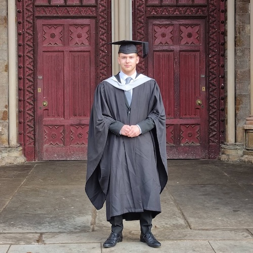 Photo of Robert standing in front of St Albans Cathedral in Graduation robes